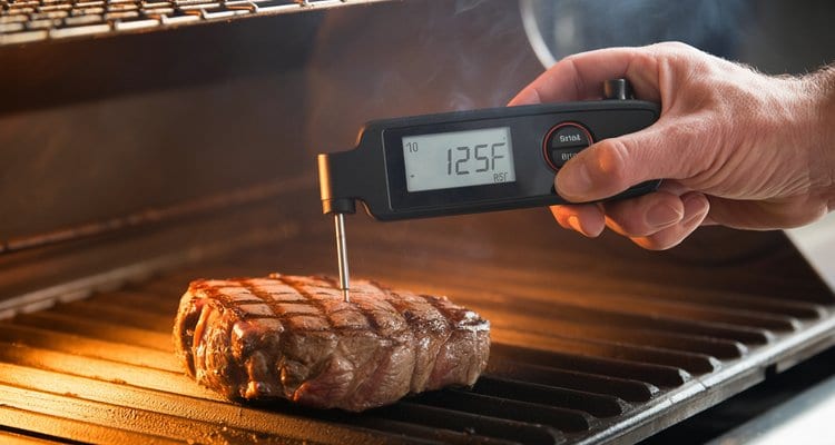 Meat thermometer side insertion technique on grilled steak Hand inserting instant-read meat thermometer horizontally into a steak on a grill showing 125°F pull-off temperature